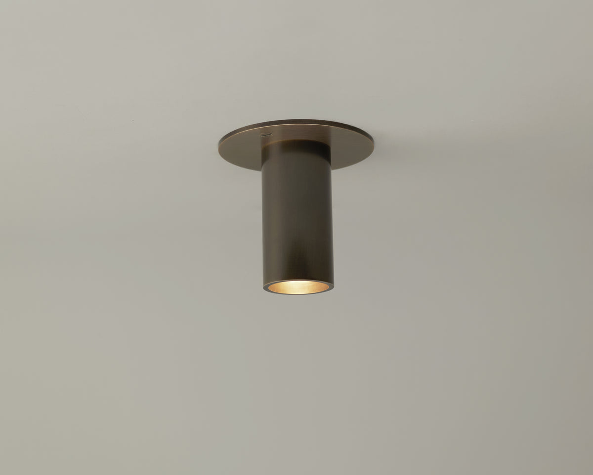 The Long Made Co. Micro Flush Spot, a bronze-colored fixture, is installed on a white ceiling and casts a gentle downward glow ideal for accent lighting.
