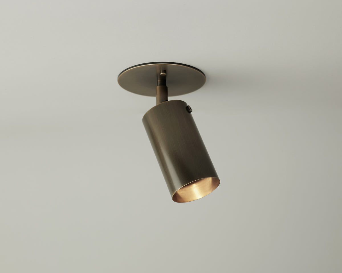 The Brass Cylinder Adjustable Spot - Top Mount by Long Made Co. is a sleek mid-century modern brass cylinder light with a round base, mounted on a white ceiling and angled downward for stylish directional lighting.