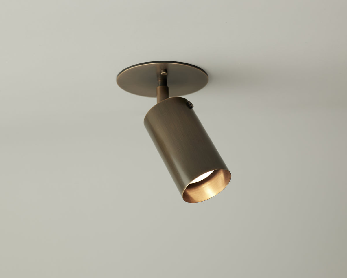 The Long Made Co. Brass Cylinder Adjustable Spot - Top Mount is a stylish, modern ceiling downlight with a cylindrical brass body on a round base, providing directional lighting against a light grey ceiling.
