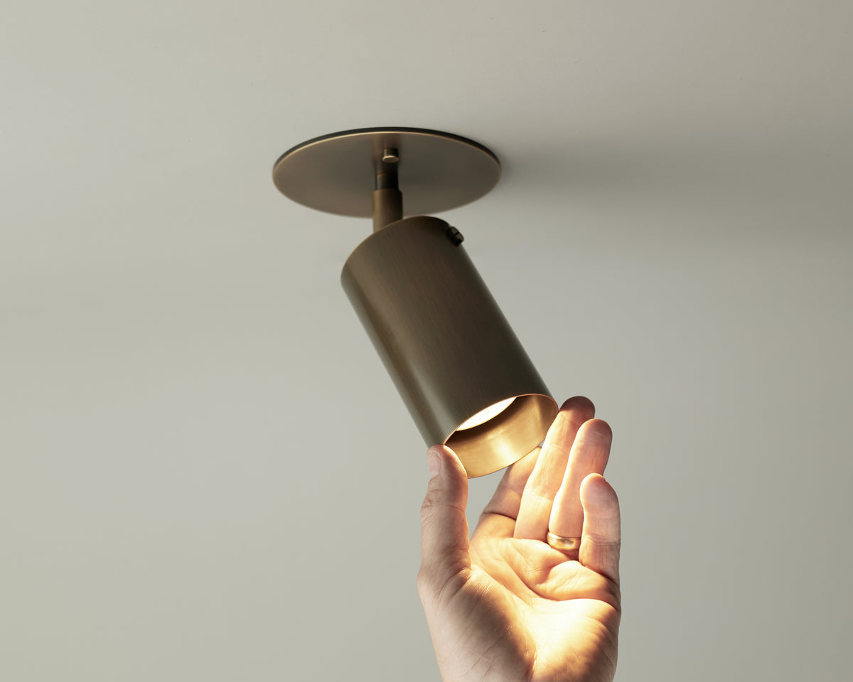 A hand with a gold ring adjusts the Long Made Co. Brass Cylinder Adjustable Spot - Top Mount, a ceiling downlight that's on and casting warm, mid-century modern style illumination.