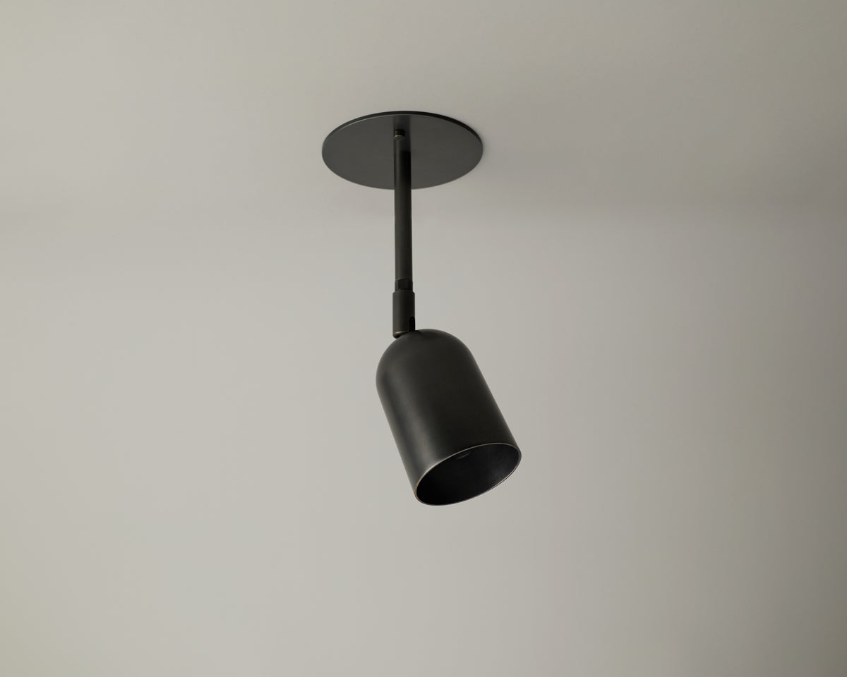 The Campana Drop Spot by Long Made Co. is a black, cylindrical ceiling spotlight fixture mounted on a white ceiling and angled slightly downward. This modern, minimalist directional downlight provides focused illumination exactly where you need it most.