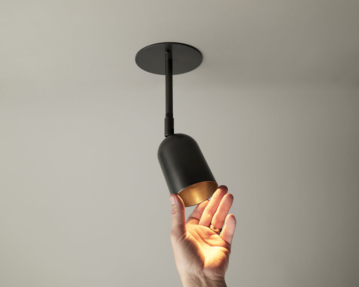 A hand reaches up toward the Long Made Co. Campana Drop Spot, a black directional ceiling downlight that is turned on, casting a warm glow over the hand and part of the fixture against a plain light-colored ceiling.