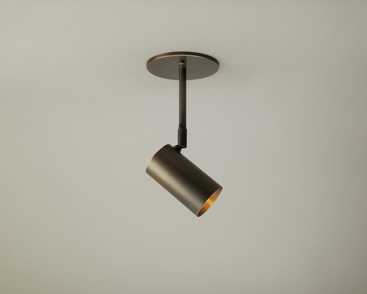 A single Brass Cylinder Drop Spot - Side Mount track light fixture by Long Made Co. is mounted on a round ceiling plate against a plain light gray ceiling. Reminiscent of mid-century modern lighting, the brass fixture features a modern, minimalist design and is angled slightly downward for focused illumination.