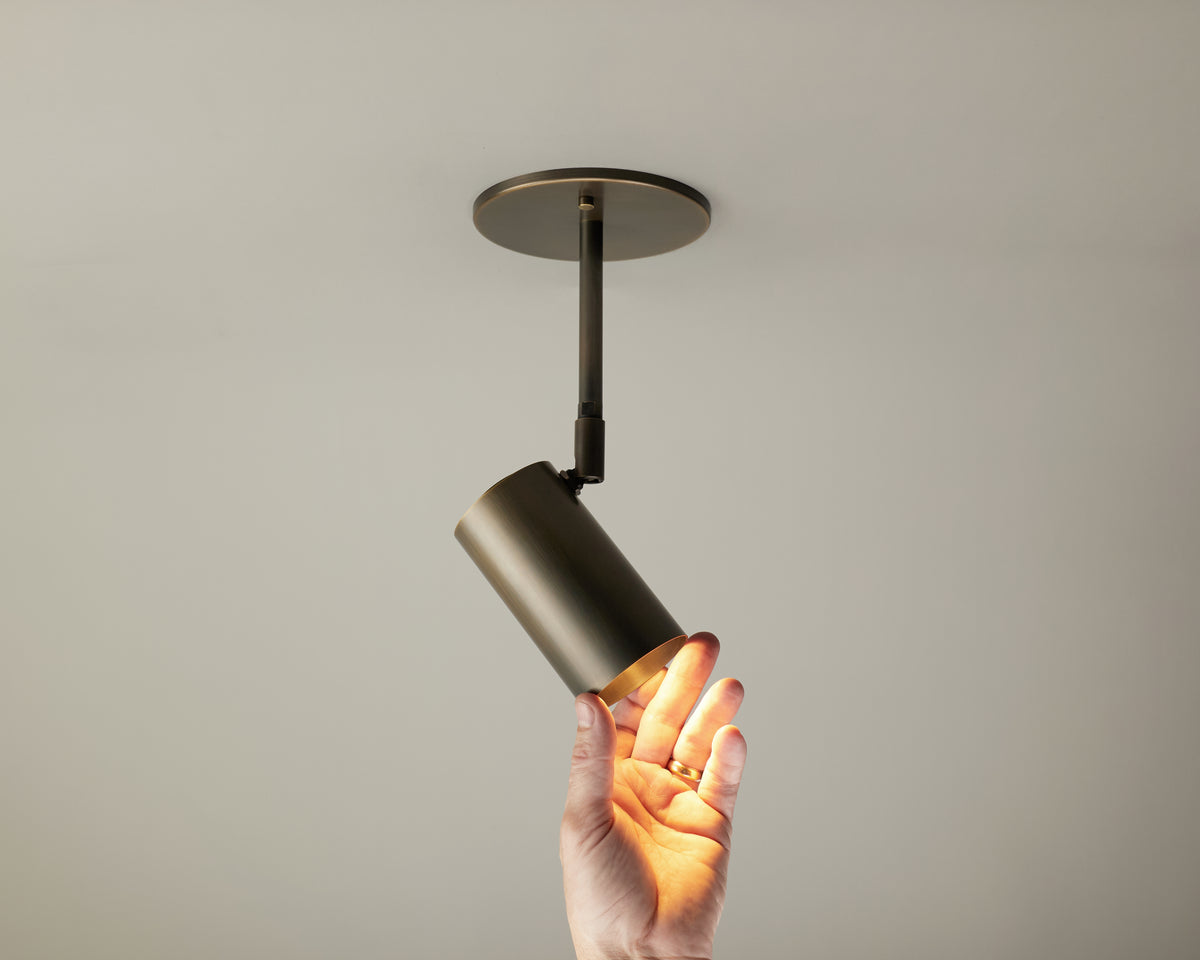 A hand touches the Brass Cylinder Drop Spot - Side Mount by Long Made Co., a ceiling-mounted cylindrical spotlight emitting a warm glow that adds an elegant touch of mid-century modern lighting to the neutral-colored room.
