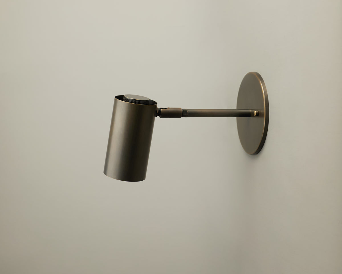 A modern Brass Cylinder Extension Wall Lamp by Long Made Co. is mounted on a plain, light-colored wall. Featuring a horizontal arm and minimalistic cylindrical design with a metallic brass finish, this handcrafted fixture brings elegant simplicity to contemporary interiors.