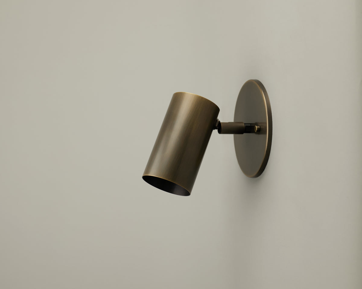The Brass Cylinder Wall Lamp by Long Made Co. is mounted on a simple, light-colored wall and angled slightly downward. This mid-century modern sconce functions as an adjustable reading light while bringing timeless elegance to your space.