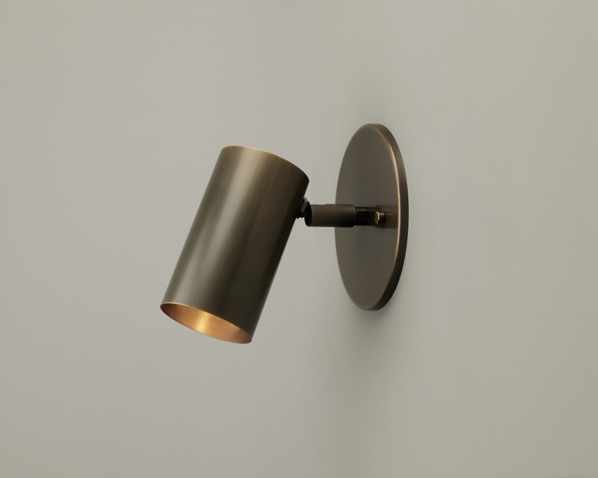 The Brass Cylinder Wall Lamp by Long Made Co. is a mid-century modern wall sconce with a solid brass finish and a cylindrical shade, mounted on a round base against a plain light-gray wall, and angled downward for use as an adjustable reading light.