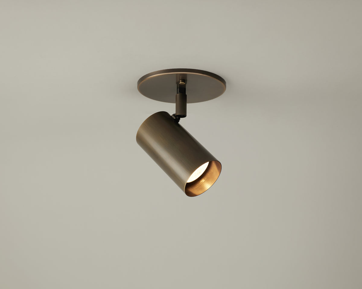 A single Brass Cylinder Adjustable Spot - Side Mount by Long Made Co. is mounted on the ceiling with a round base, featuring a brass-colored cylindrical form designed as an adjustable directional downlight that casts light downward at an angle against a plain, light background.