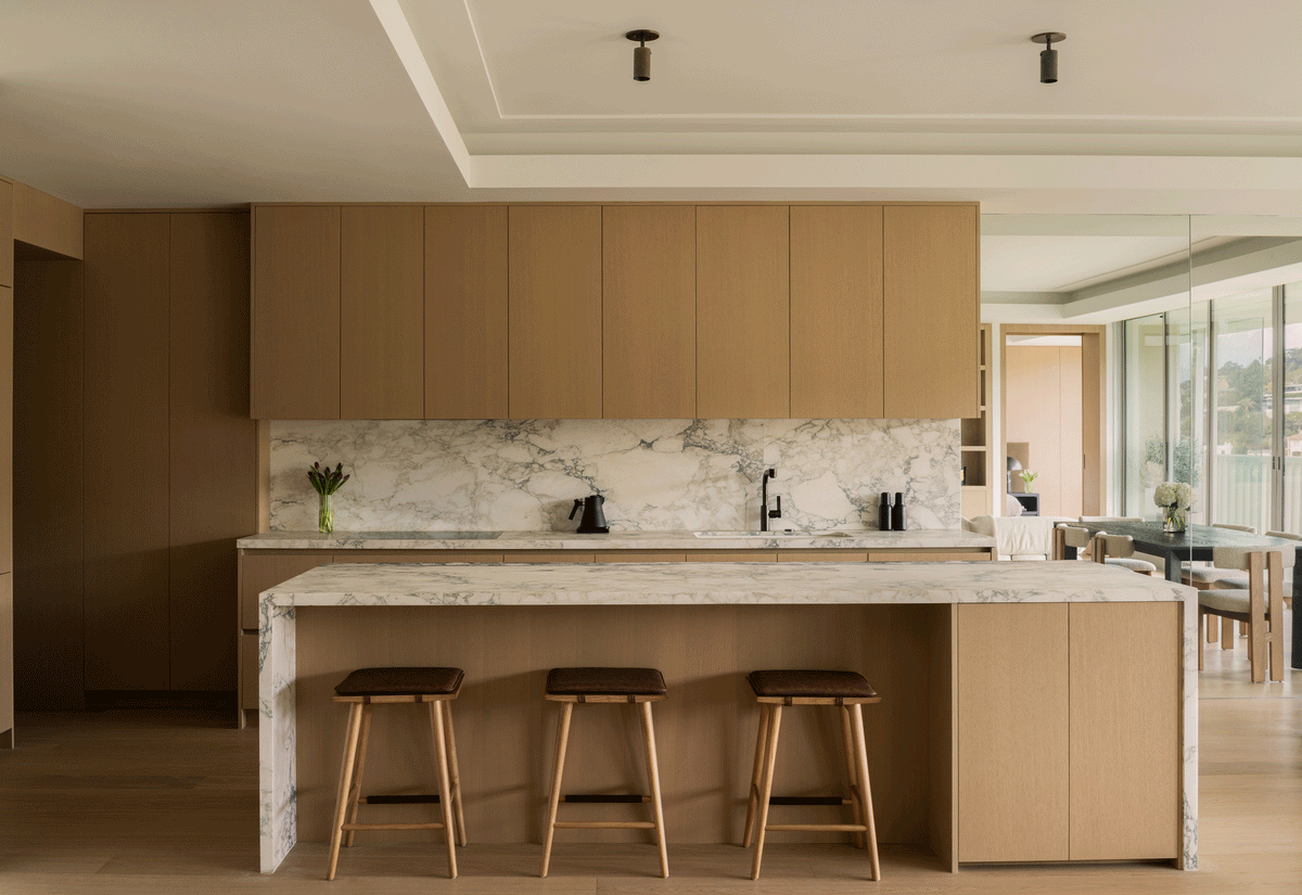 Modern kitchen with light wood cabinets, marble surfaces, and three wooden bar stools. Large windows and a dining area feature the Brass Cylinder Adjustable Spot - Top Mount by Long Made Co. for mid-century modern lighting accents.