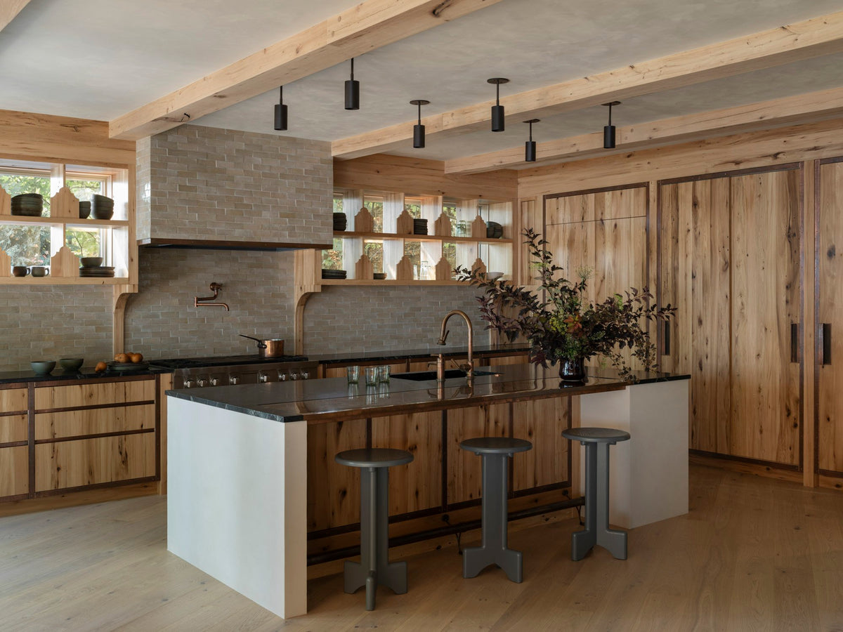 A modern rustic kitchen features light wood cabinets, exposed beams, and a central island with three gray stools. Highland Drop Spot brass spotlights by Long Made Co. illuminate the island. Open shelves display pottery and a vase with branches.