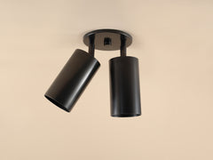 The Highland Double Spot by Long Made Co. is a modern ceiling light featuring two cylindrical black spotlights on a round base, showcased against a beige background with minimalist design.