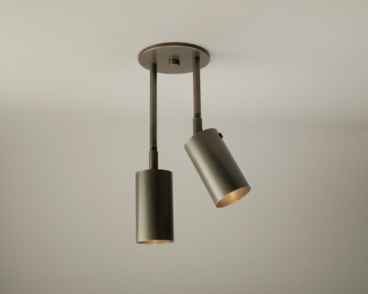 Discover the Long Made Co. Brass Cylinder Double Drop Spot—a modern ceiling light fixture featuring two cylindrical, adjustable spotlights in a matte metallic finish, mounted on a circular base against a plain white ceiling. This distinctive mid-century modern piece offers versatile directional lighting for any space.