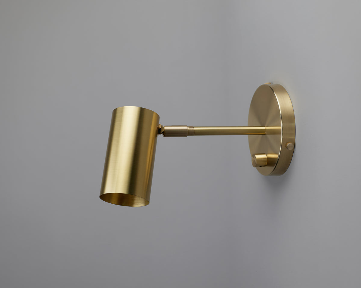 The Brass Cylinder Extension Wall Lamp by Long Made Co. is mounted on a light gray wall. This mid-century modern fixture showcases a sleek, minimalist design with a round base and an adjustable arm, making it ideal as an adjustable reading light for any contemporary space.