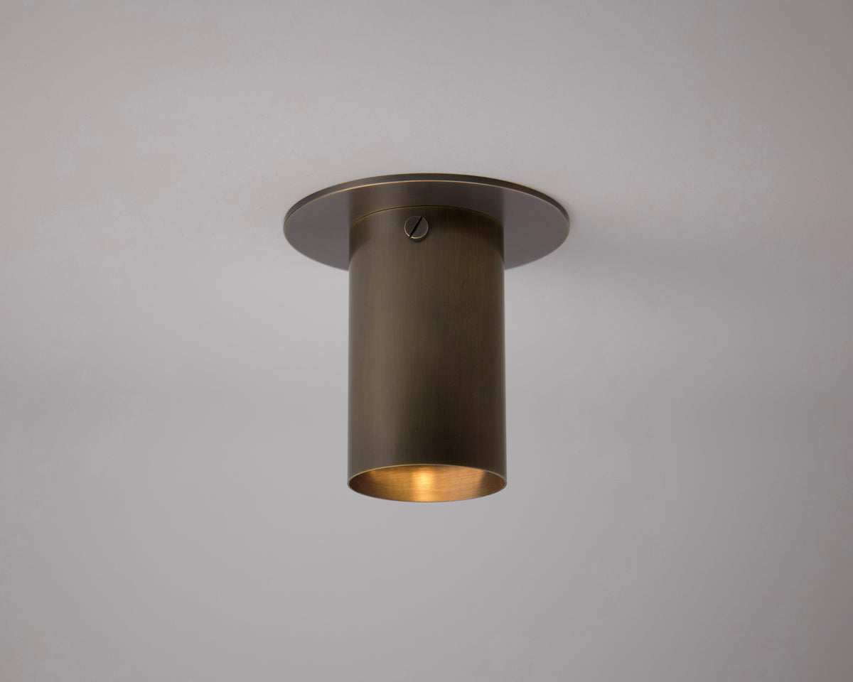 The Long Made Co. Brass Cylinder Flush Spot, featuring solid brass construction and a brushed metal finish, mounts flush to a white ceiling and casts a soft, warm downward glow.