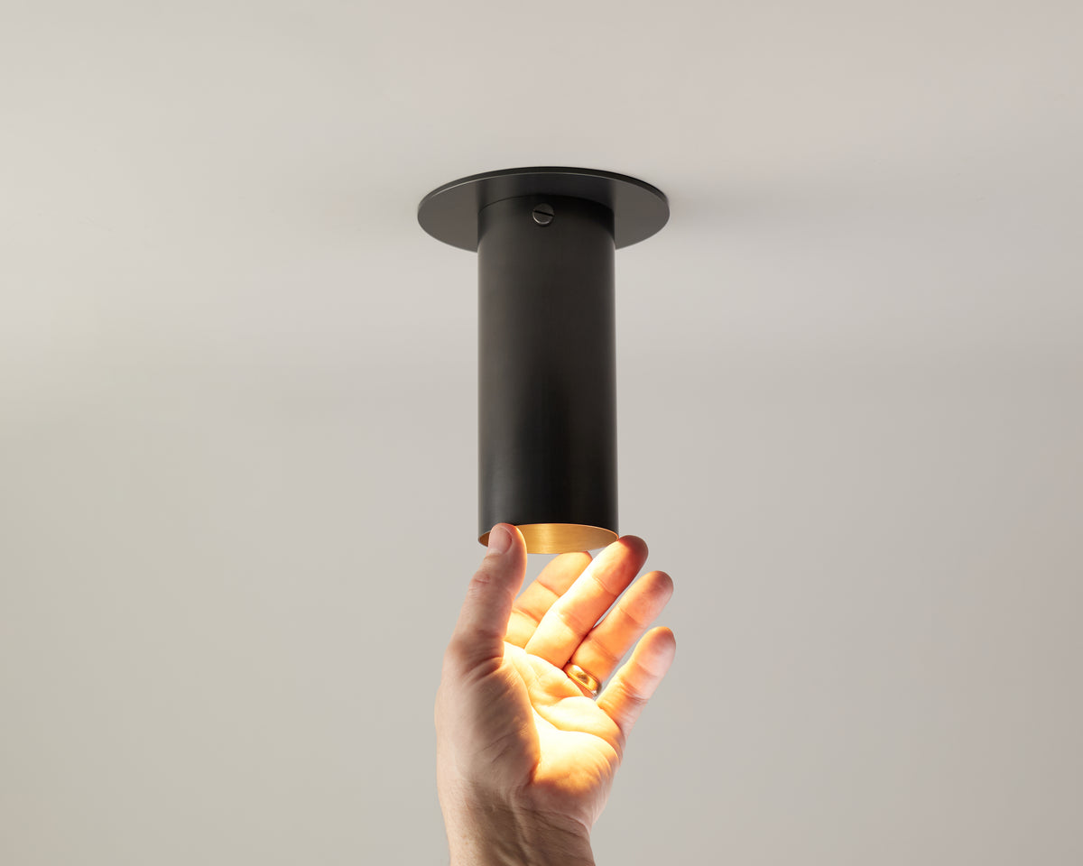A hand reaches up toward the Brass Cylinder Flush Spot with Long Shade by Long Made Co., its sleek black cylindrical form casting a warm, mid-century modern glow across the palm against a plain white background.