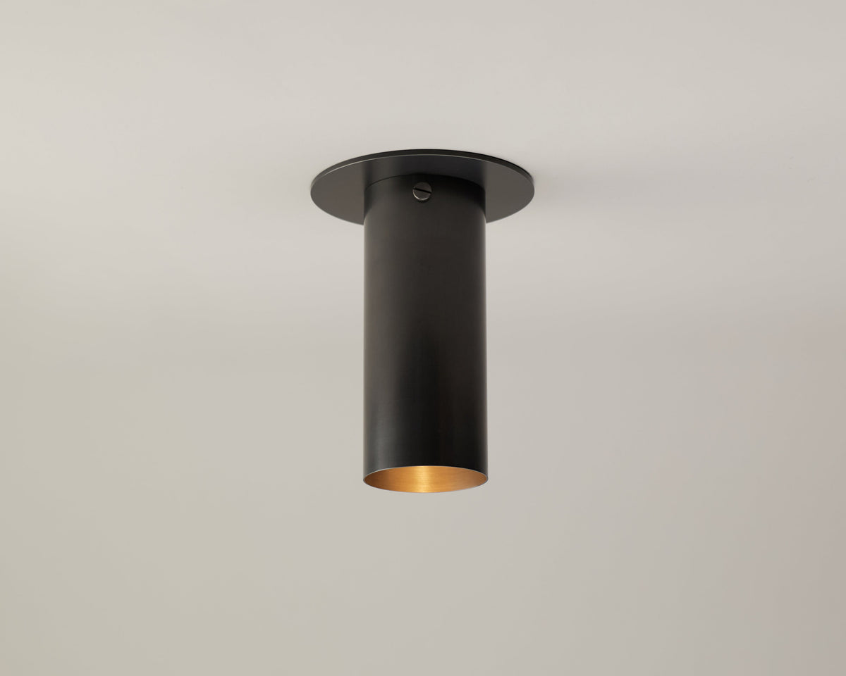 A matte black Brass Cylinder Flush Spot with a long shade by Long Made Co., inspired by mid-century modern lighting, is mounted on a plain white ceiling and emits light from its open bottom.