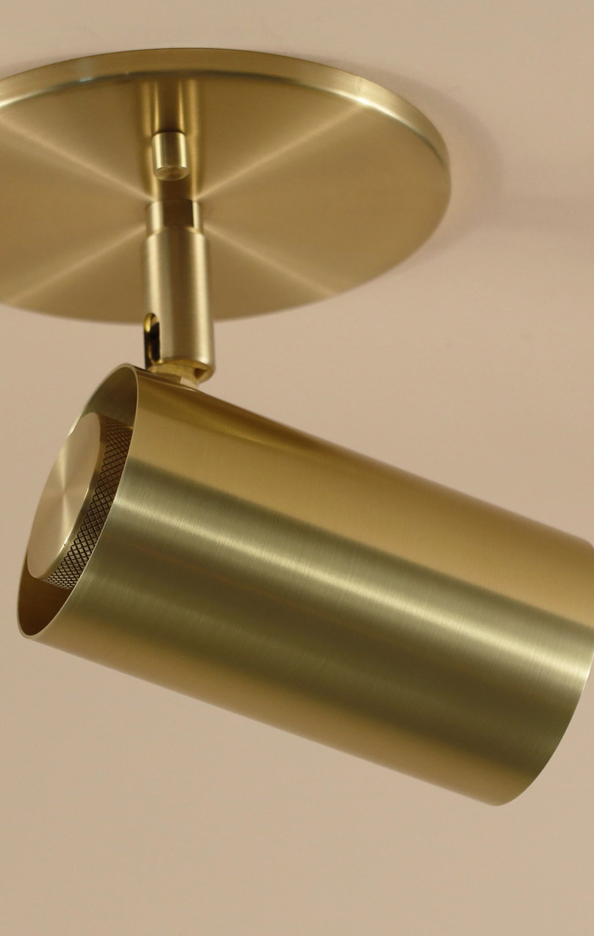 A close-up of the Brass Cylinder Adjustable Spot - Side Mount by Long Made Co., with its cylindrical solid brass body on a round base, showcases sleek mid-century modern style against a light ceiling.