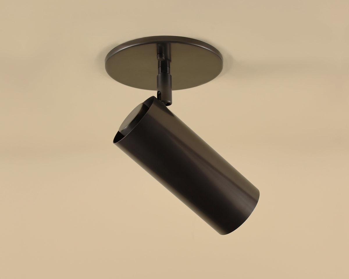 The Brass Cylinder Adjustable Spot - Side Mount w/ Long Shade by Long Made Co. is a modern black cylindrical track light, angled downward and mounted on a beige ceiling, evoking mid-century modern style.
