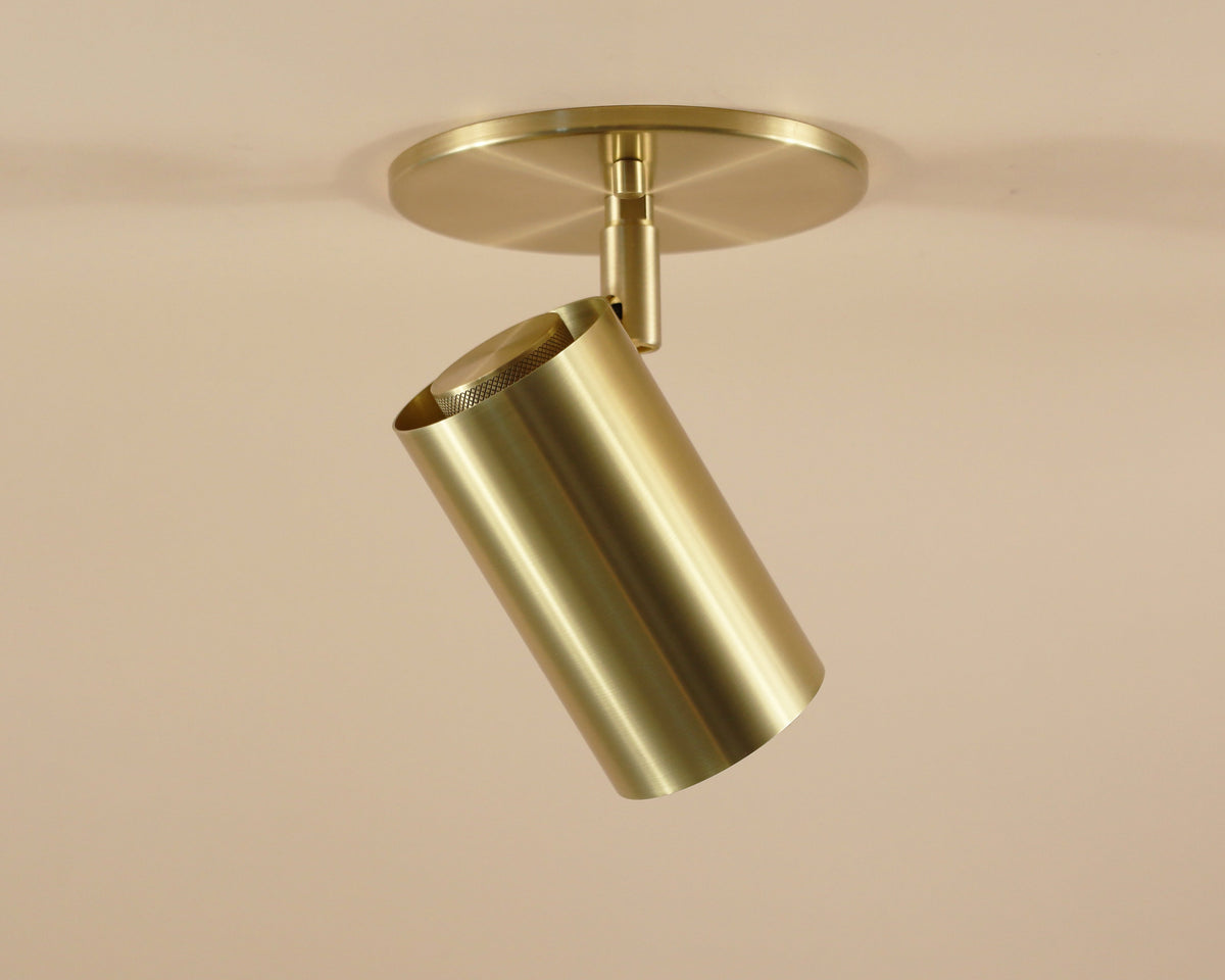 The Long Made Co. Brass Cylinder Adjustable Spot - Side Mount features a mid-century modern brushed gold finish with a round base, installed on a light-colored ceiling and angled downward for customizable lighting.