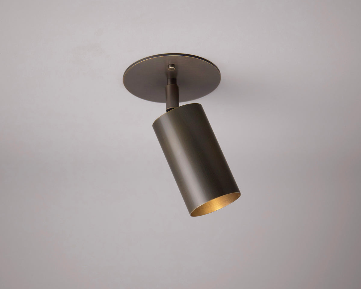 The Brass Cylinder Adjustable Spot - Top Mount by Long Made Co. is a modern, cylindrical fixture with a dark metallic finish and gold interior, angled downward against a plain light gray background.