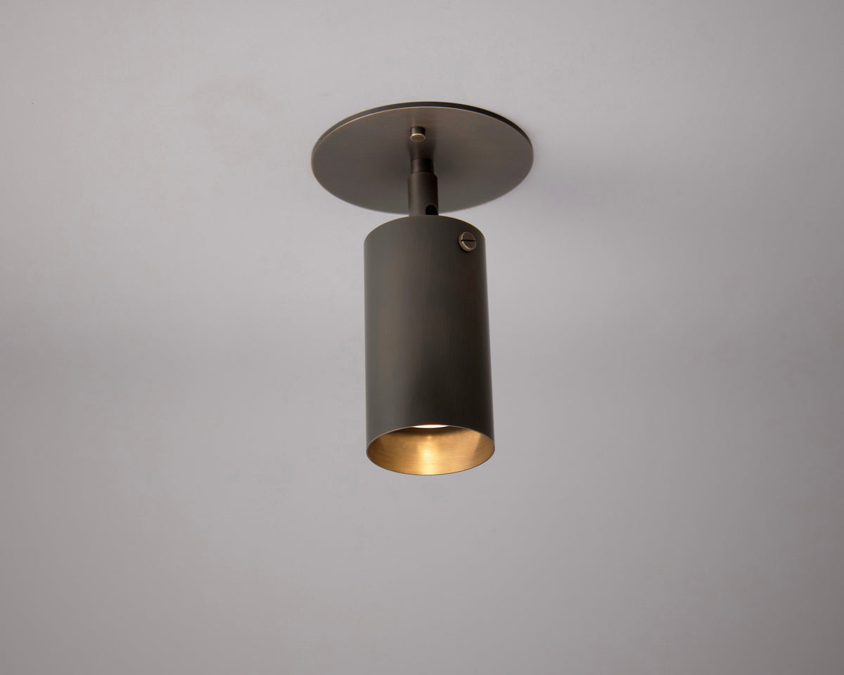 The Long Made Co. Brass Cylinder Adjustable Spot - Top Mount is a cylindrical metallic fixture with a warm glow from its open bottom, mounted on a plain light ceiling.