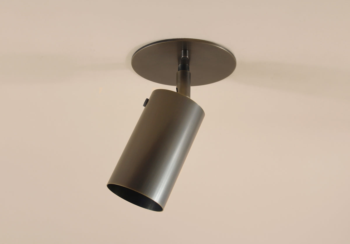 The Brass Cylinder Adjustable Spot - Top Mount by Long Made Co. is a modern ceiling fixture with a cylindrical brass design on a circular base, angled downward for adjustable lighting and timeless style.
