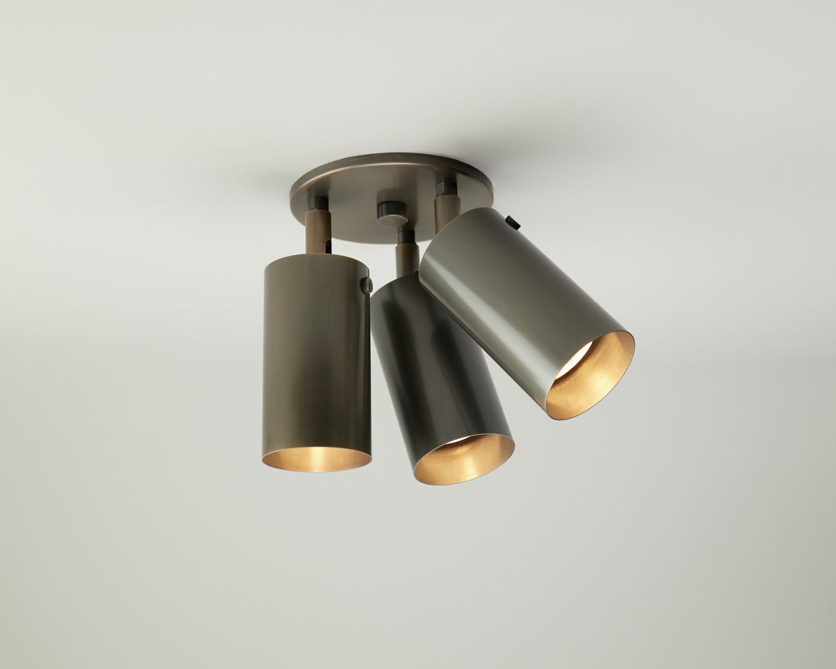 The Brass Cylinder Triple Spot by Long Made Co. is a modern ceiling light fixture featuring three adjustable cylindrical spotlights, each pointing in a different direction, mounted on a round base against a white ceiling.