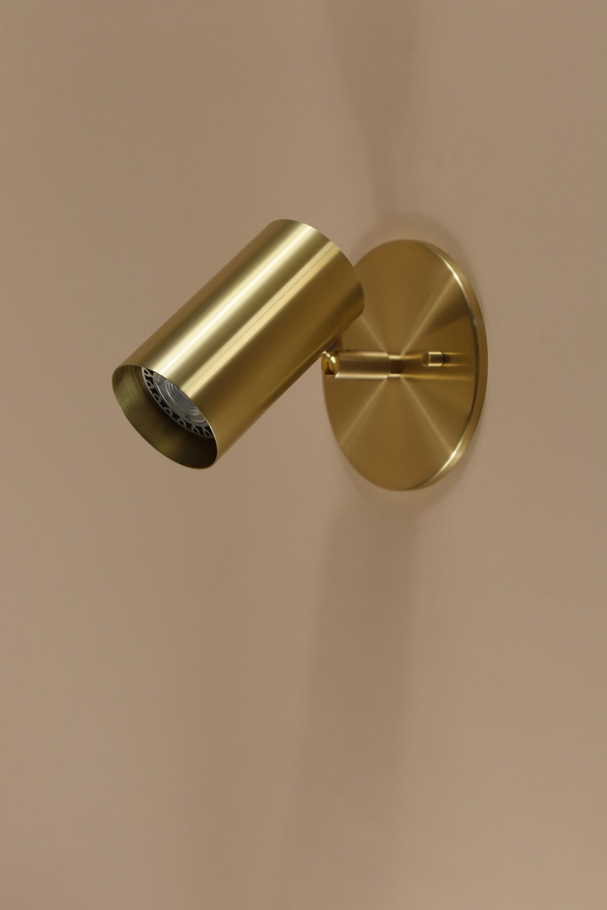 The Brass Cylinder Wall Lamp by Long Made Co. is a modern, cylindrical sconce with a round base, mounted on a light beige wall. Its sleek, minimalist brass design is angled slightly downward for stylish illumination.