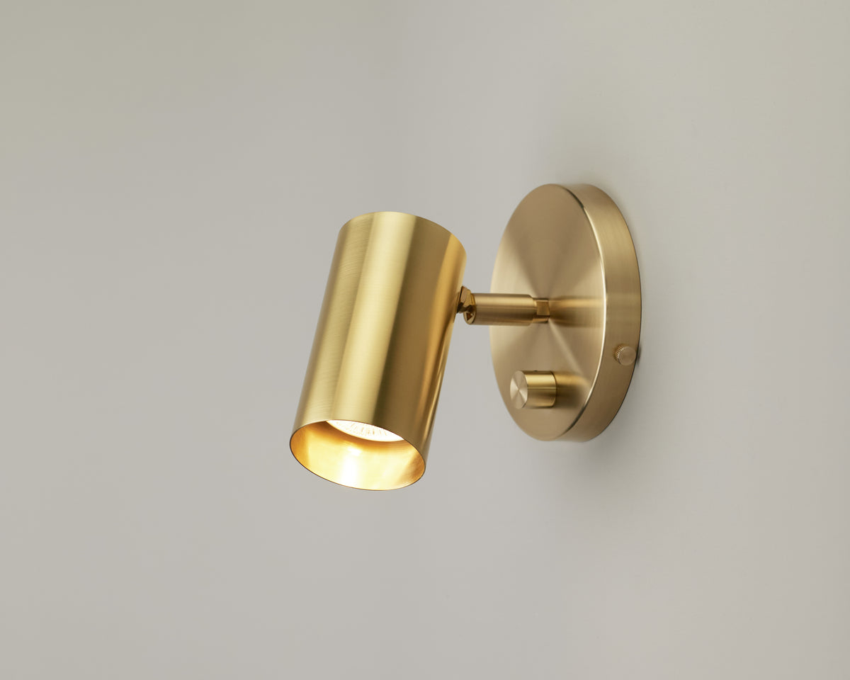The Brass Cylinder Wall Lamp by Long Made Co. features a modern solid brass design with a cylindrical shade mounted on a round base, casting a soft, warm glow against a light-colored wall.