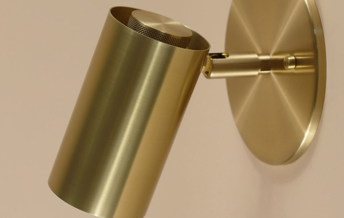 A close-up of the Brass Cylinder Wall Lamp by Long Made Co., featuring a modern brushed gold brass finish, cylindrical shade, and circular base, mounted on a light-colored wall.