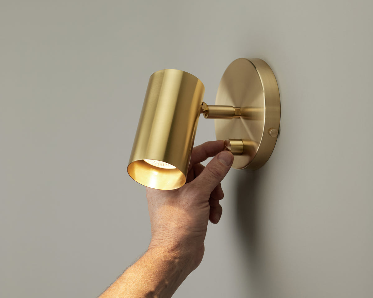 A hand adjusts the Long Made Co. Brass Cylinder Wall Lamp, a modern gold-colored sconce, on a plain, light-colored wall.