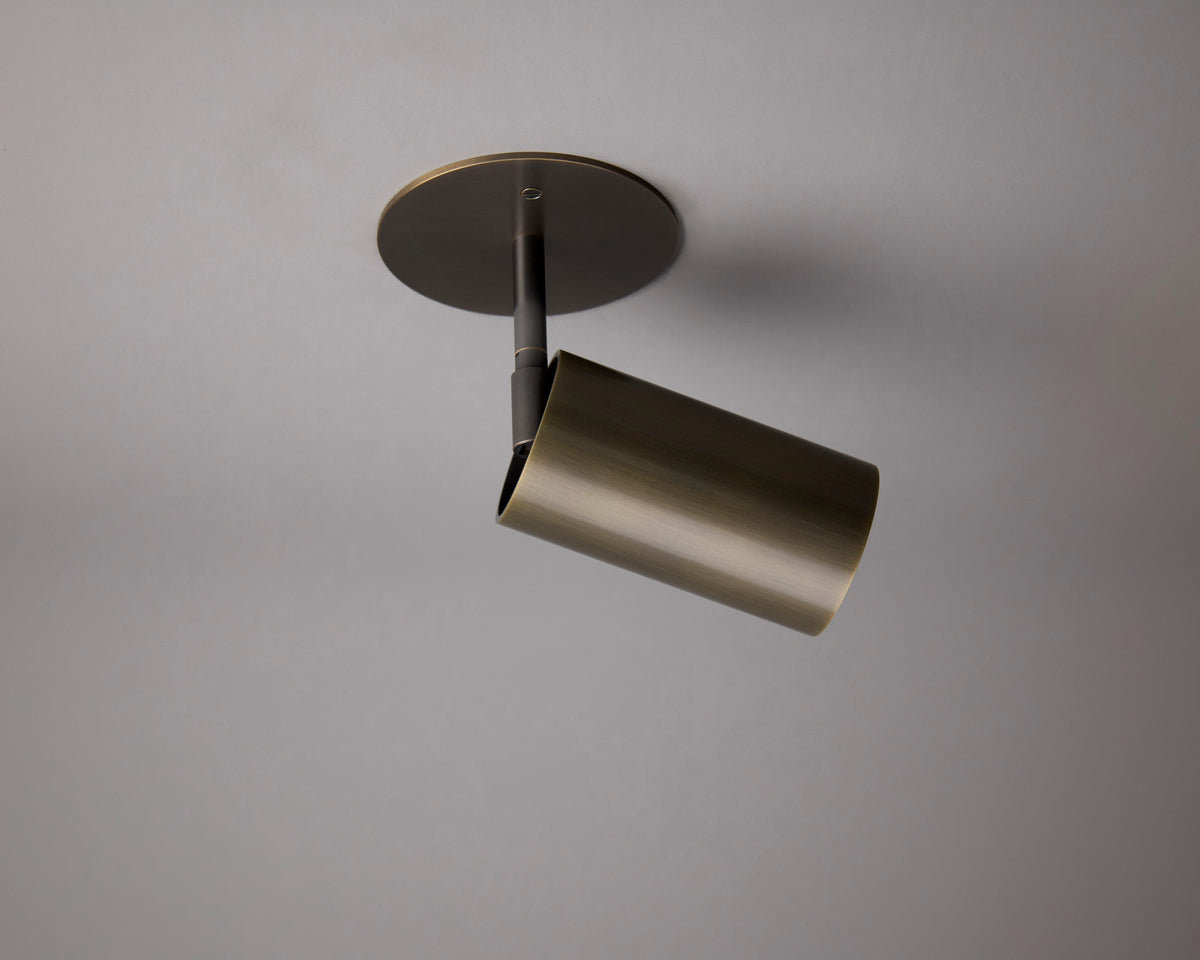 The Beau Laiton Drop Monopoint by Long Made Co. is a bronze ceiling spotlight in a minimalist brass style, mounted on a round base against a light gray ceiling, with the light angled downward.
