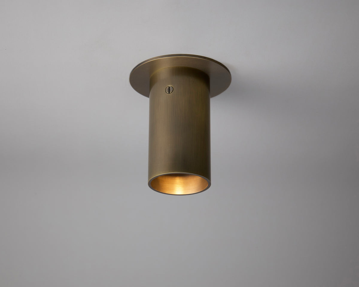 The Beau Laiton Flush Spot by Long Made Co. is a solid brass cylindrical spotlight mounted on a plain white ceiling, providing a warm downward glow with its minimalist design.