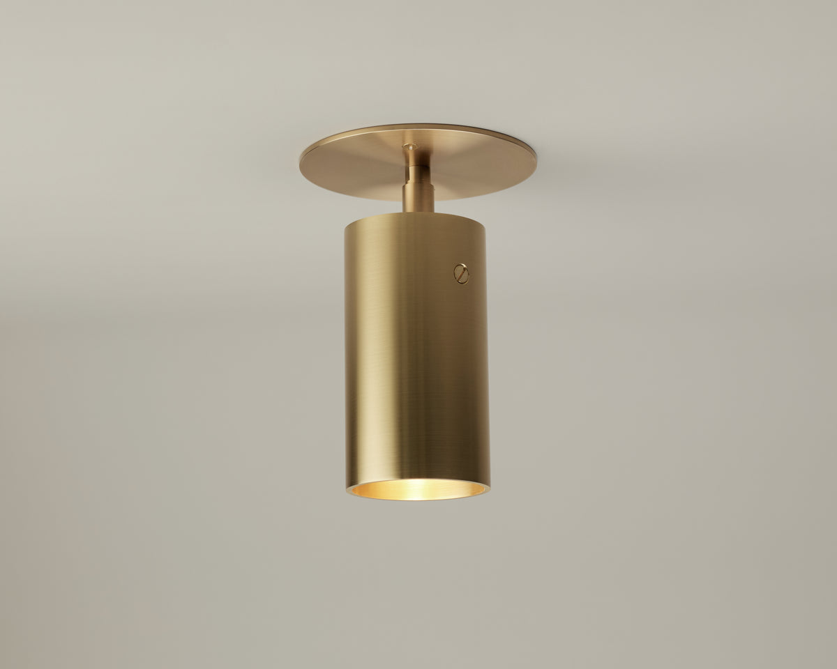 The Beau Laiton Monopoint by Long Made Co. is a minimalist brushed brass cylindrical ceiling light fixture with a round base, mounted on a plain white ceiling and casting a soft downward glow—an understated elegant lighting piece that enhances any space.
