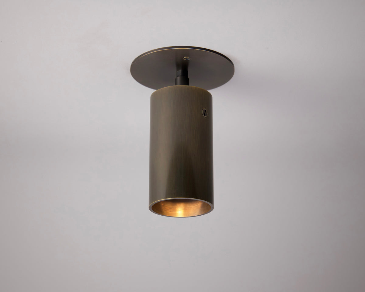 The Beau Laiton Monopoint by Long Made Co. is a modern, cylindrical bronze ceiling fixture mounted on a white ceiling, casting a warm downward glow—a refined homage to mid-century lighting design.