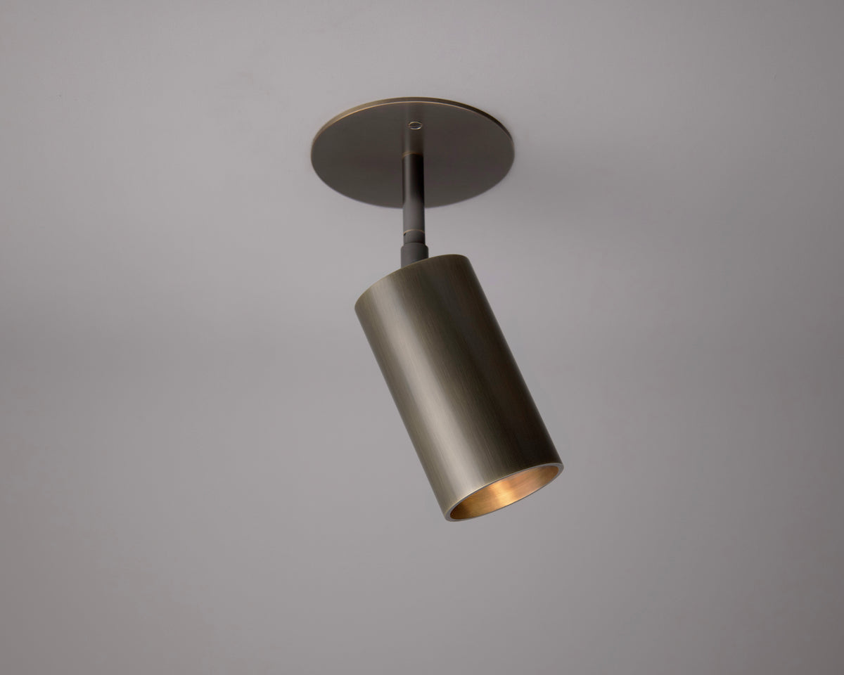 The Beau Laiton Drop Monopoint by Long Made Co. is a modern cylindrical ceiling fixture with a brushed brass finish, mounted on a round base and angled downward against a plain light gray ceiling.