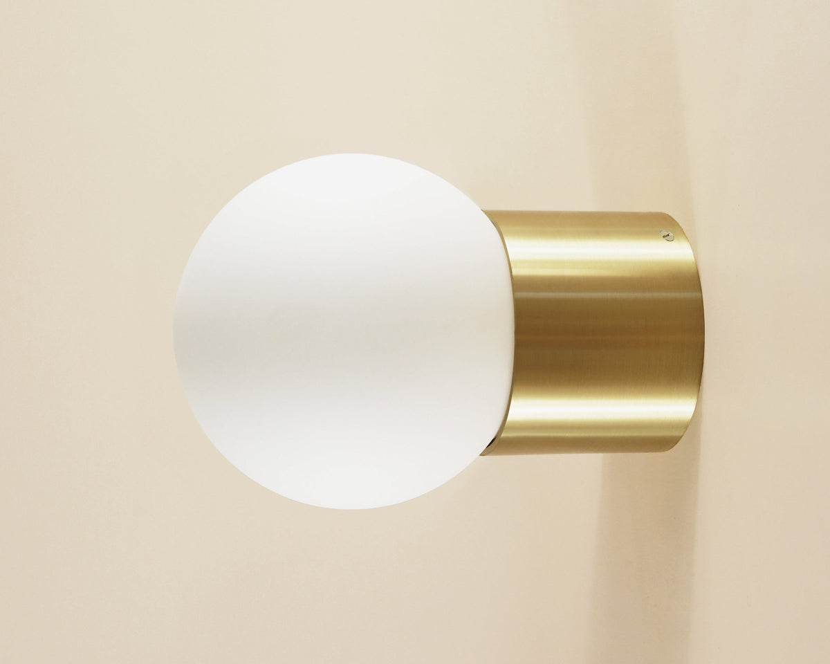 Wall-mounted light fixture with gold finish and white glass shade on a beige background