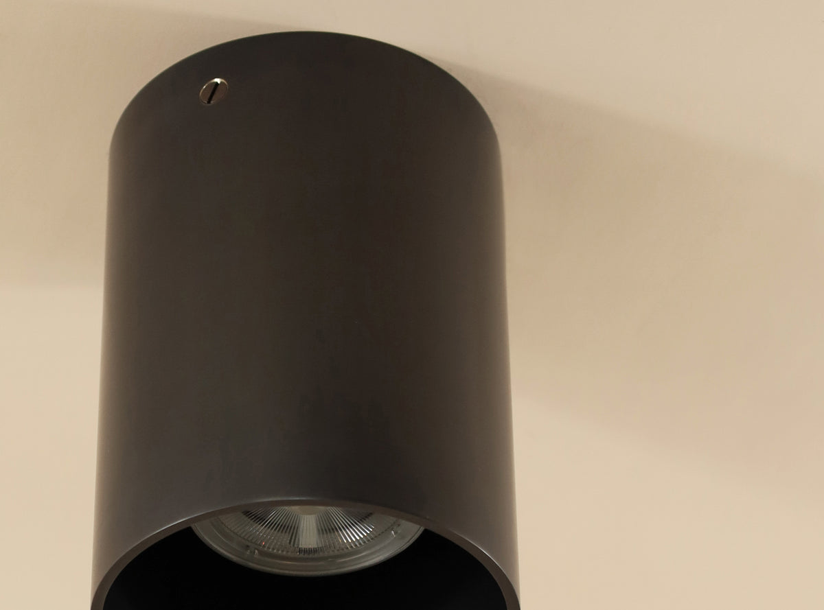 A close-up of the Long Made Co. Beau Laiton Trimless Downlight, a black cylindrical ceiling fixture with minimalist brass accents, mounted on a beige ceiling. The light bulb is partially visible inside the fixture.