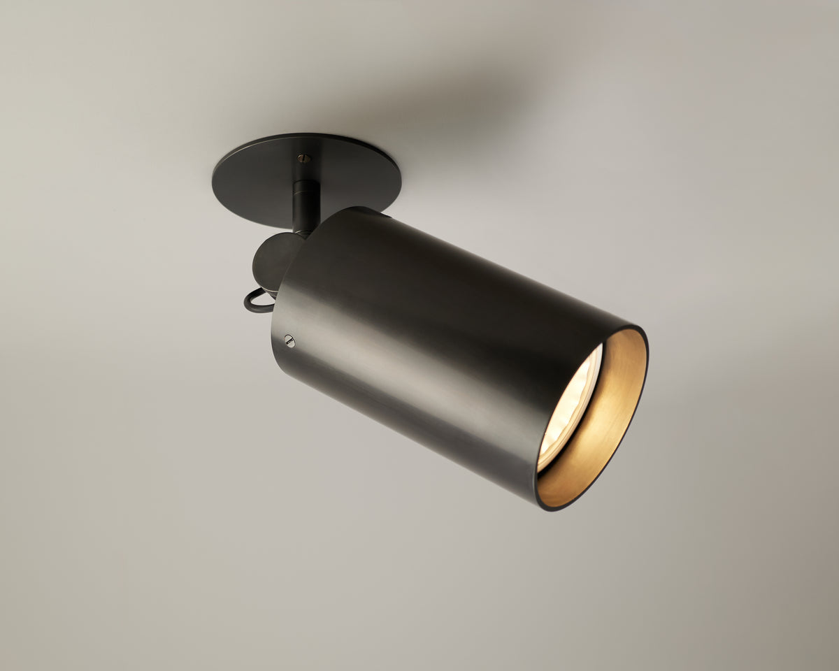A modern black cylindrical ceiling spotlight fixture—the Beau Laiton - XL Adjustable Spot by Long Made Co.—evokes the adjustable spot light design found in Lutron Ketra S30 collections. It is mounted on a white ceiling, with the light turned on and casting a warm glow.