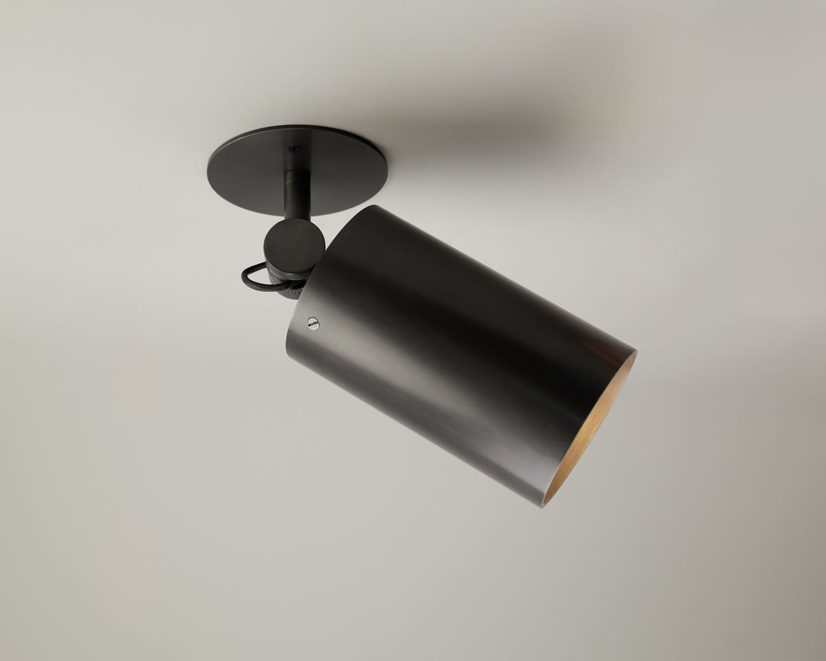 The Beau Laiton - XL Adjustable Spot by Long Made Co. is a black cylindrical fixture, inspired by solid brass lighting design, mounted on a round base and angled downward against a plain white ceiling.