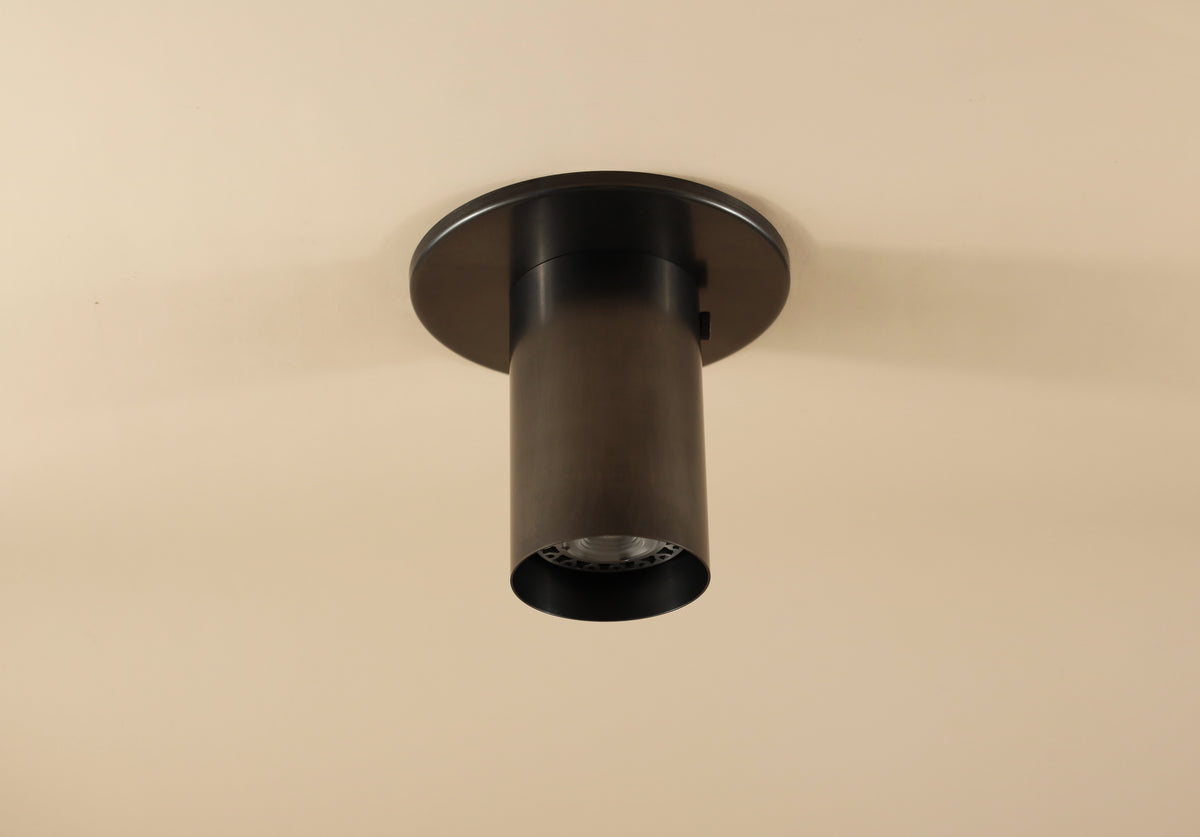 The Brass Cylinder Flush Spot by Long Made Co. is a modern, cylindrical black recessed ceiling light fixture inspired by mid-century design, shown here mounted on a beige ceiling.