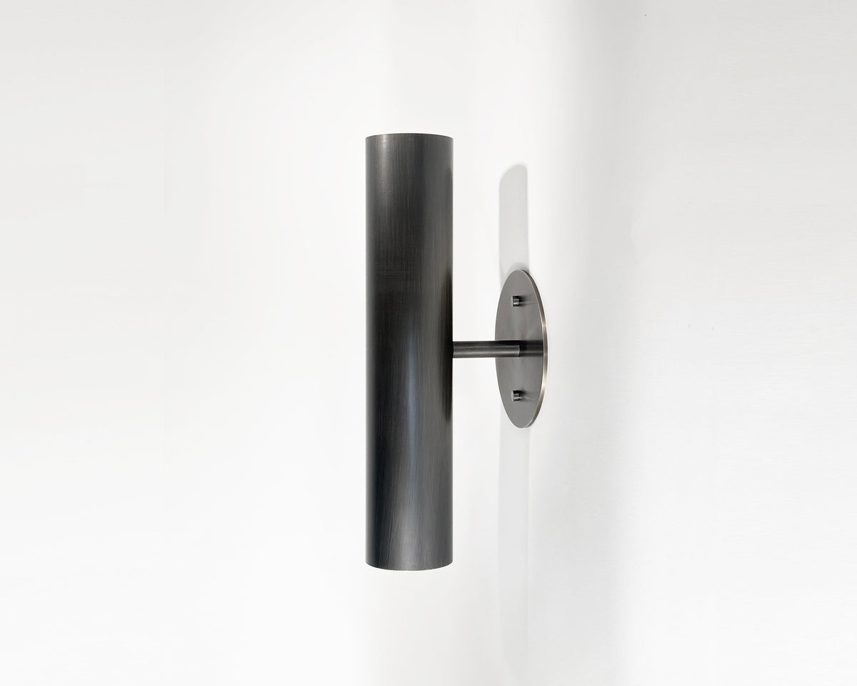 The Brass Cylinder Up-Down Sconce by Long Made Co. features a modern, brushed brass cylindrical design, mounted vertically on a white wall for a sleek mid-century modern lighting touch.