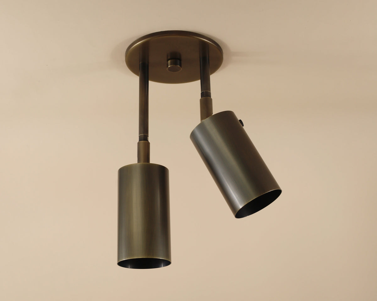 The Long Made Co. Brass Cylinder Double Drop Spot is a modern ceiling fixture with two brass spotlights on a round base, angled for versatile lighting—ideal for fans of mid-century modern style.