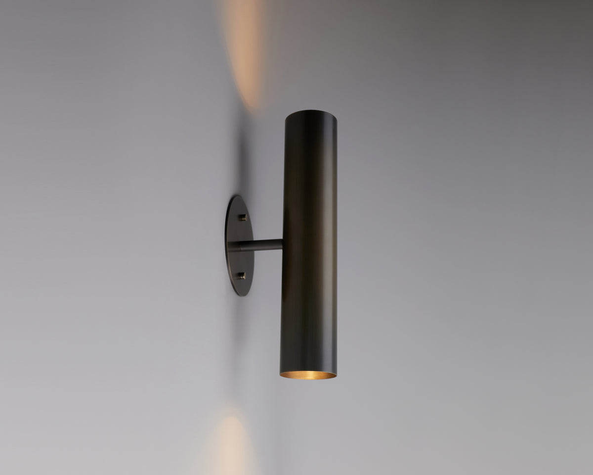 A modern Brass Cylinder Up-Down Sconce by Long Made Co. is mounted vertically, emitting light from both the top and bottom and casting a soft glow on the smooth gray wall—an effortless nod to mid-century modern lighting.