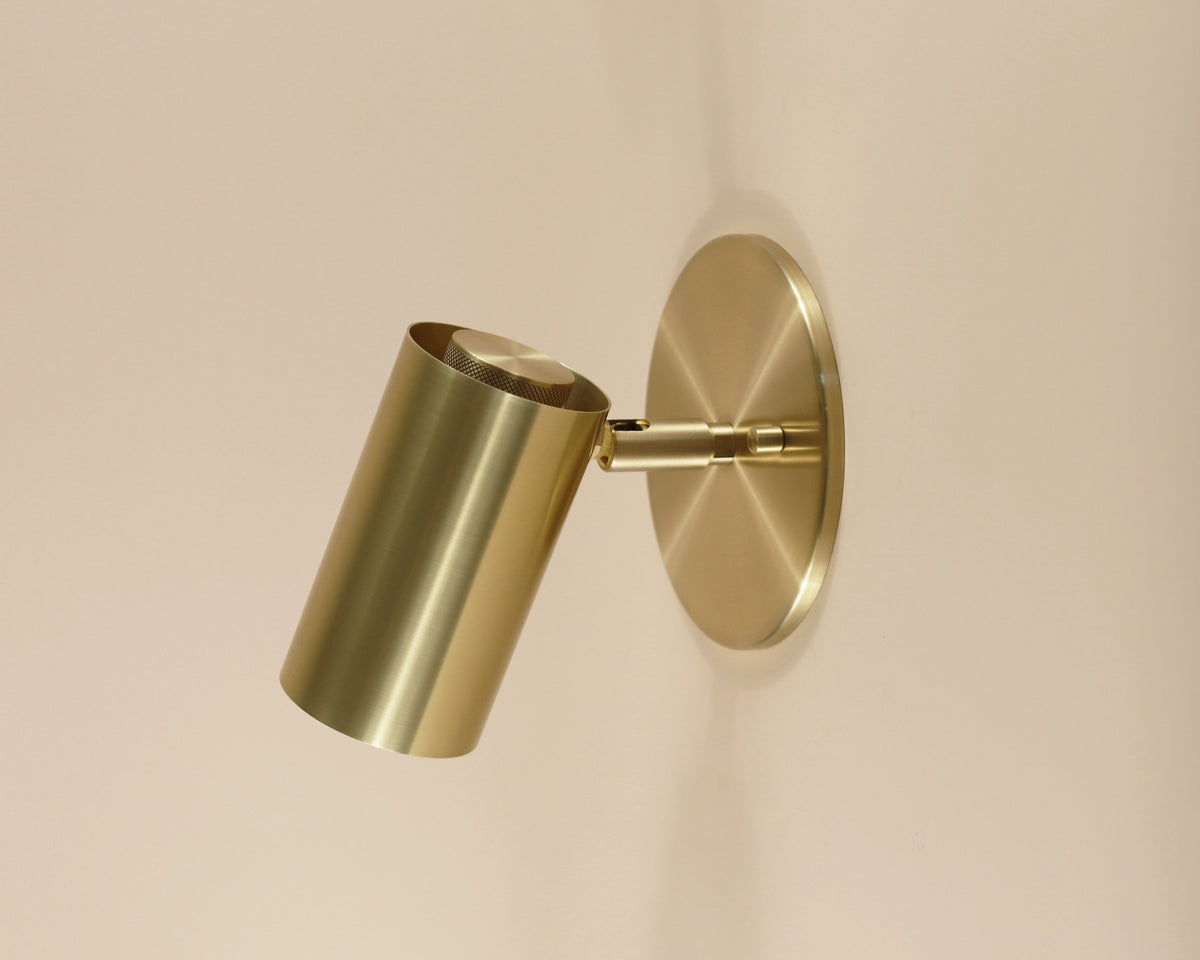 The Brass Cylinder Wall Lamp by Long Made Co. is a modern brass sconce with a cylindrical shade and round base, blending mid-century style and function for any light-colored wall.