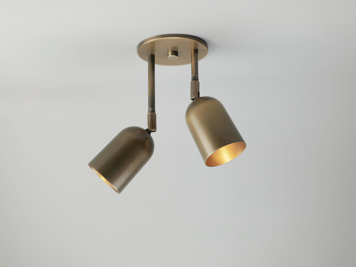 The Campana Double Drop Spot by Long Made Co. is a brushed brass ceiling light fixture featuring two adjustable cylindrical shades. Mounted against a plain white ceiling, one shade is angled lower to emit focused light. Crafted from solid brass, this versatile piece functions beautifully as a directional spot, delivering both timeless style and concentrated illumination.
