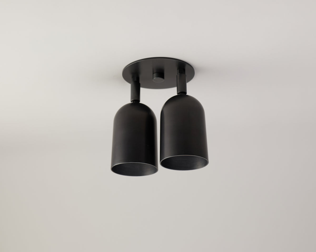 The Campana Double Spot by Long Made Co. is a modern black ceiling light fixture featuring two cylindrical shades on a round base, set against a plain white ceiling and evoking the elegance of a mid-century spotlight.