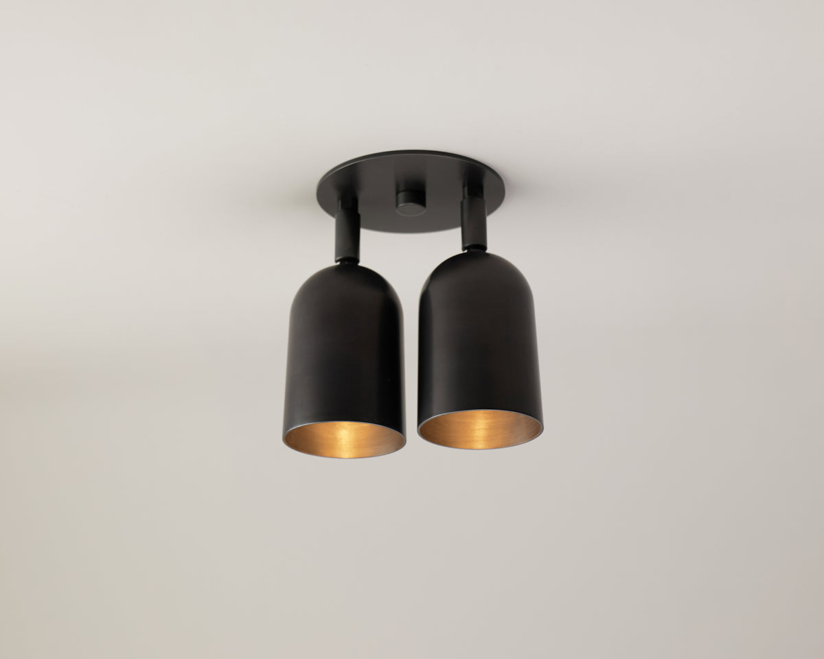 The Campana Double / Triple Spot by Long Made Co. is a modern black ceiling light fixture featuring two cylindrical downward-facing bulbs mounted on a round base, bringing a touch of mid-century spotlight style to a plain white ceiling.