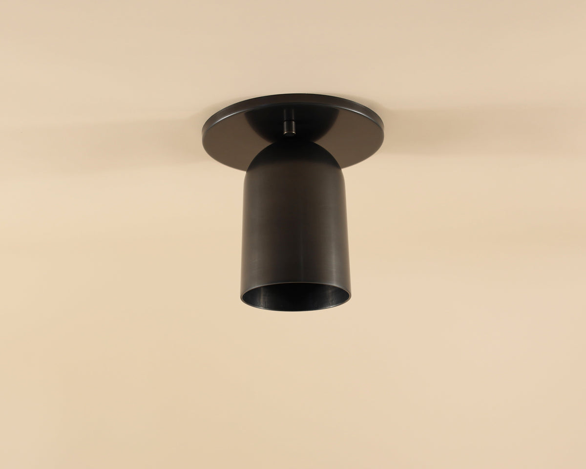 The Campana Flush Spot by Long Made Co. is a modern black cylindrical ceiling downlight with solid brass construction, stylishly mounted on a beige ceiling as an elegant alternative to recessed can lights.
