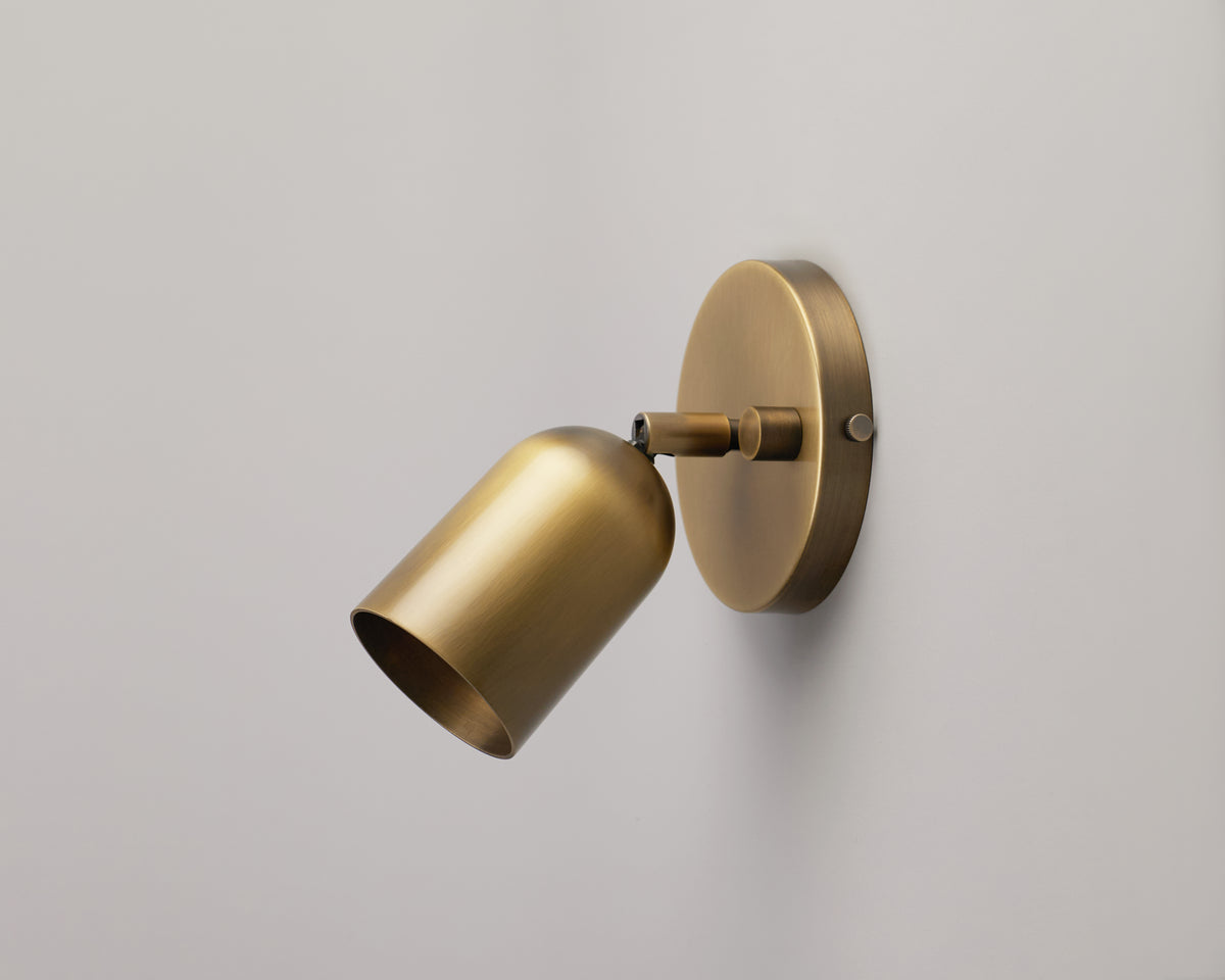 The Campana Wall Lamp by Long Made Co. features a modern solid brass design with a cylindrical shade, mounted on a circular base against a plain light-colored wall, highlighting the appeal of handmade lighting in contemporary interiors.